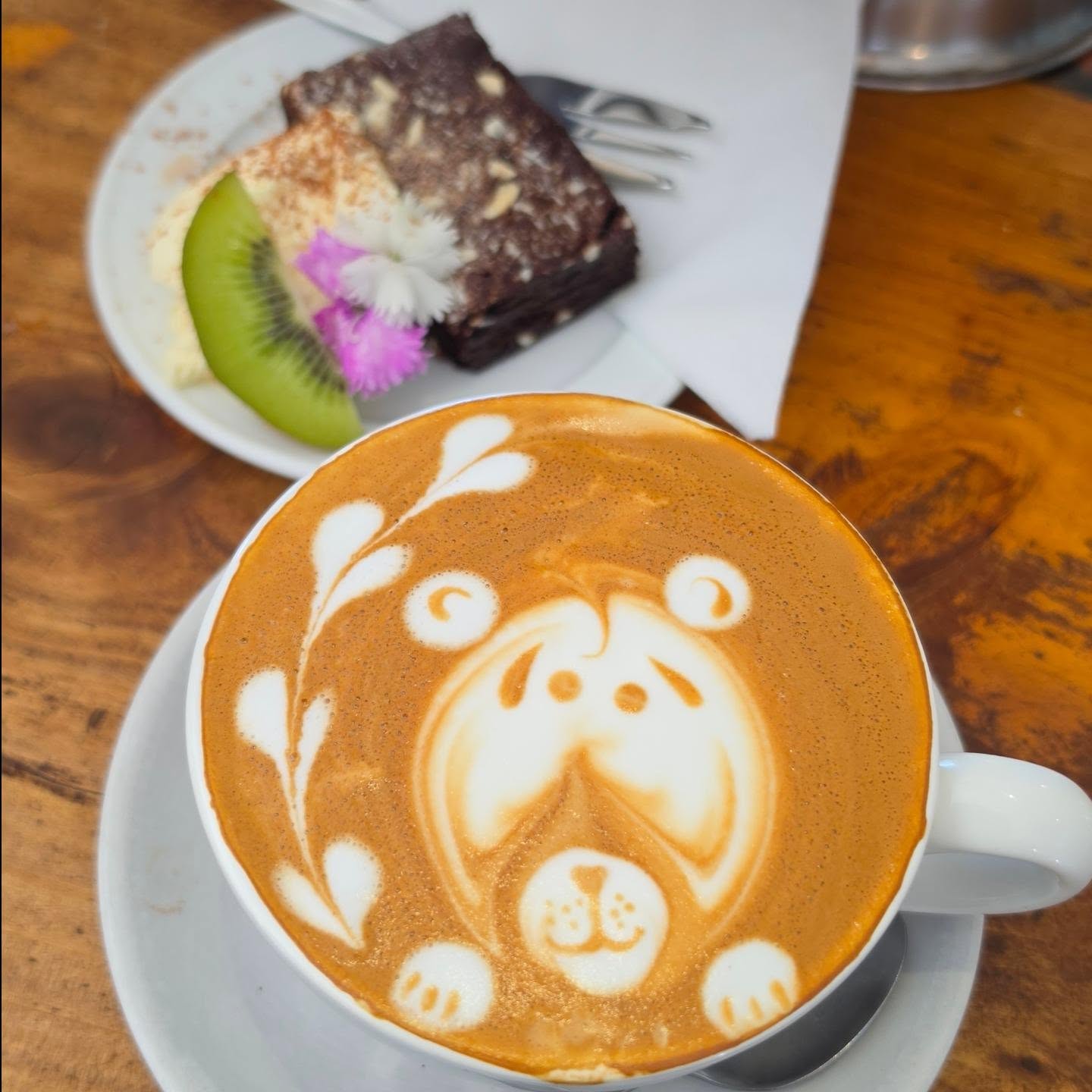 A bear made of latte art in a white cup beside a slice of cake with kiwifruit and an edible flower, on a worn timber table