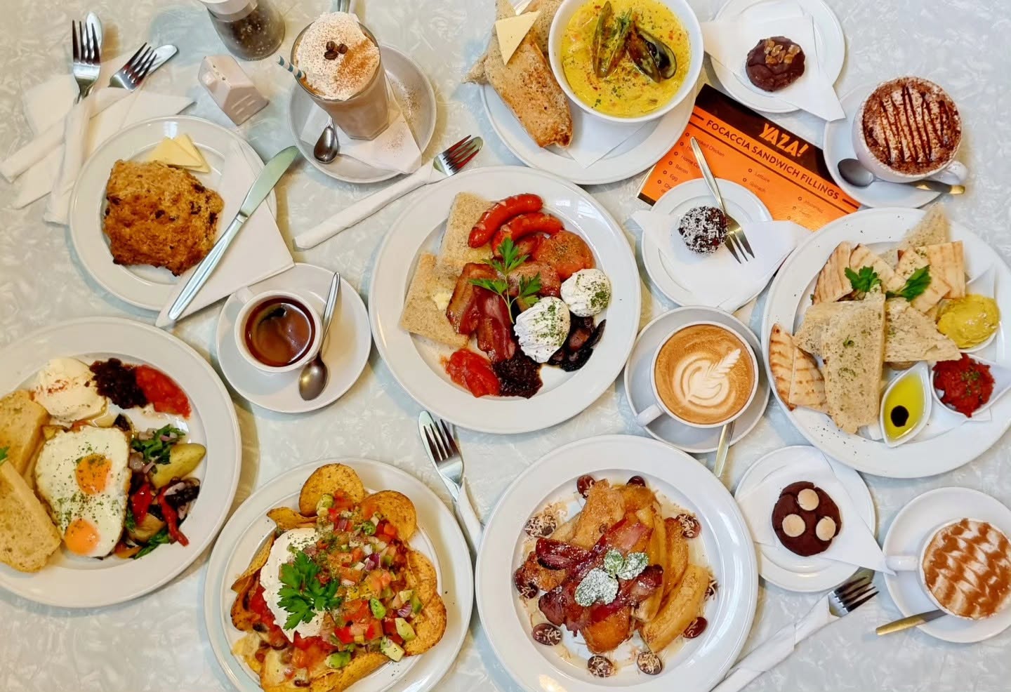 A table of dishes seen from above: a big breakfast, nachos, French toast, a mezze plate, a scone and coffees